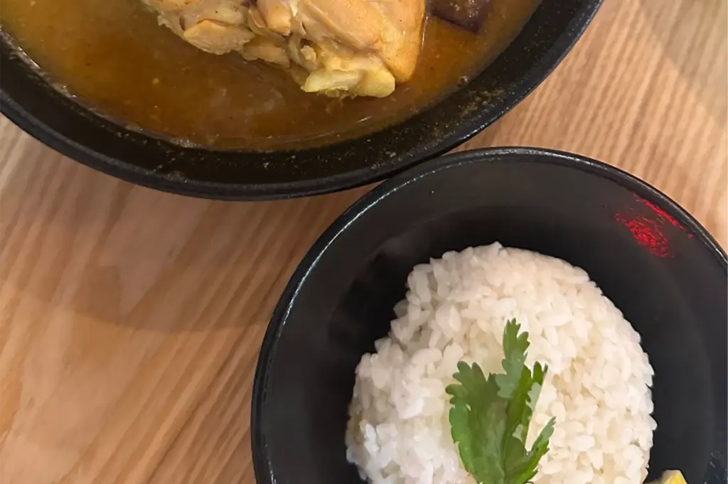 A top-down angle shows a black bowl of steamed white rice topped with a fresh sprig of cilantro, placed beside a larger bowl containing rich curry broth and tender chicken. The dish rests on a light wooden tabletop, creating a warm, inviting meal setup.