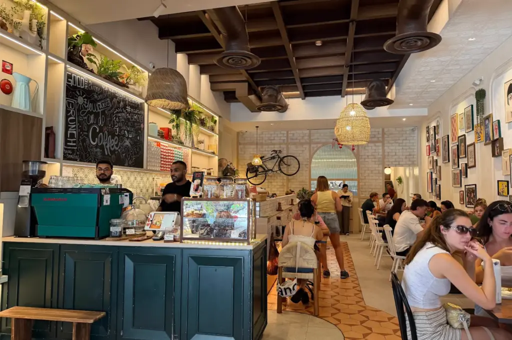 Patrons dine inside a cozy, bustling cafe featuring a dark green coffee counter with an espresso machine and pastry display case. A bicycle mounted on a white brick back wall, wicker pendant lights, chalkboard signage, and framed art add to the eclectically decorated interior.
