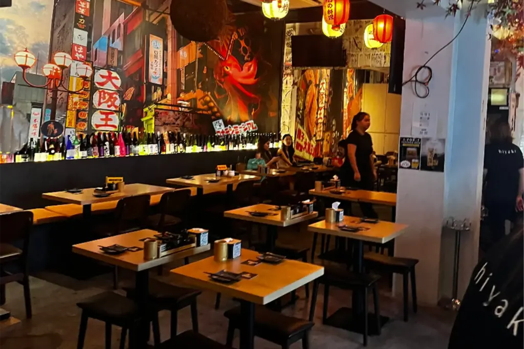 An eye-level shot captures the warm interior of a Japanese izakaya, featuring neat rows of wooden dining tables with dark stools. The space is dominated by a vibrant city-themed wall mural with a giant red octopus, accented by a long line of sake bottles and glowing paper lanterns hanging from the ceiling.