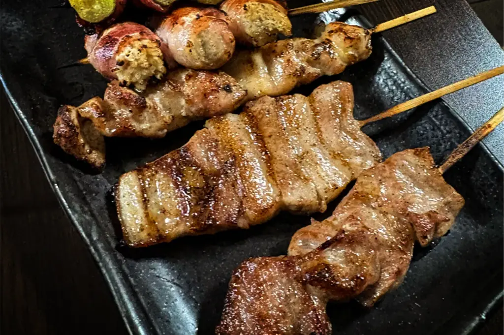 A close-up, angled shot highlights a selection of grilled Japanese yakitori skewers, including thinly sliced pork belly showing distinct grill marks, arranged on a textured black plate. In the background, wrapped skewers stuffed with a textured filling rest alongside the main cuts of meat on a dark table surface.