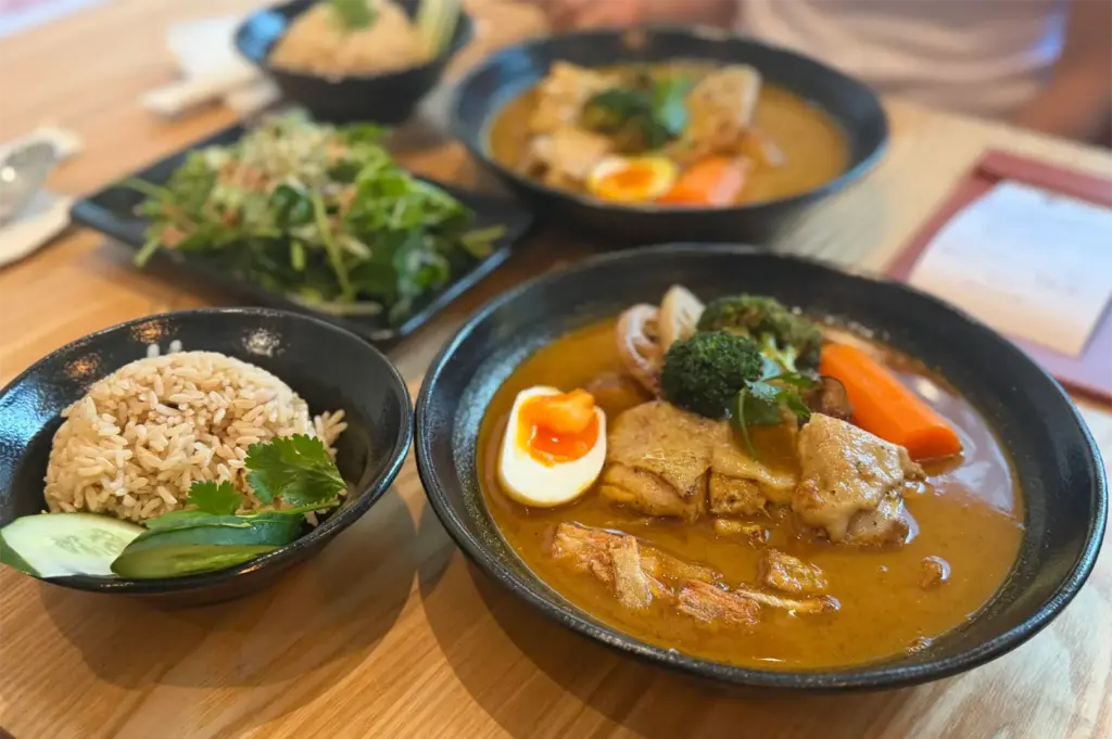 A generous meal spread features dark bowls of rich Japanese soup curry with tender chicken, soft-boiled eggs, and roasted vegetables alongside side bowls of seasoned rice garnished with cucumber and cilantro. A fresh green salad sits in the background on the light wood table, completing the vibrant table setting.