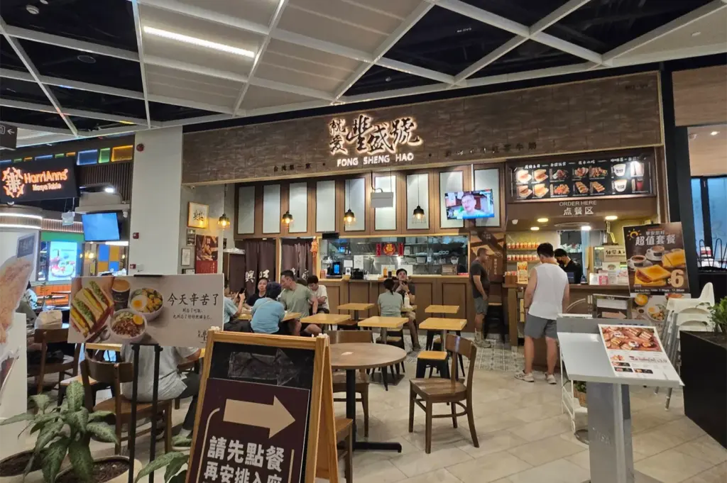 Customers sit at wooden tables inside Fong Sheng Hao, a Taiwanese eatery located within a brightly lit indoor food hall. Promotional menu stands and directional signs guide customers near the front order counter beneath a wood-paneled facade.