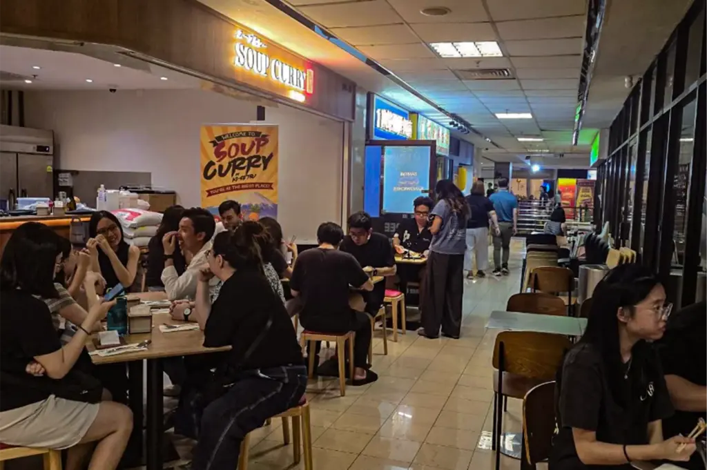 A bustling, open-front restaurant in a shopping corridor is filled with diners seated at wooden tables, enjoying their meals under a warm illuminated "Soup Curry" sign. A promotional poster stands near the entrance as patrons interact and dine along the tiled hallway.
