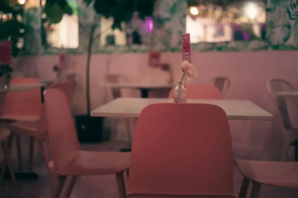 A serene, empty cafe dining space features pastel pink chairs set against white tables, with a single flower in a glass vase gracing the foreground table. Warm ambient lighting, soft pink walls, and lush green indoor foliage create a cozy, intimate atmosphere.