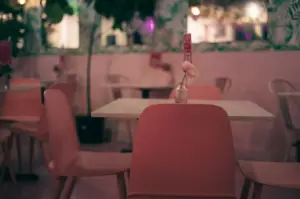 A serene, empty cafe dining space features pastel pink chairs set against white tables, with a single flower in a glass vase gracing the foreground table. Warm ambient lighting, soft pink walls, and lush green indoor foliage create a cozy, intimate atmosphere.