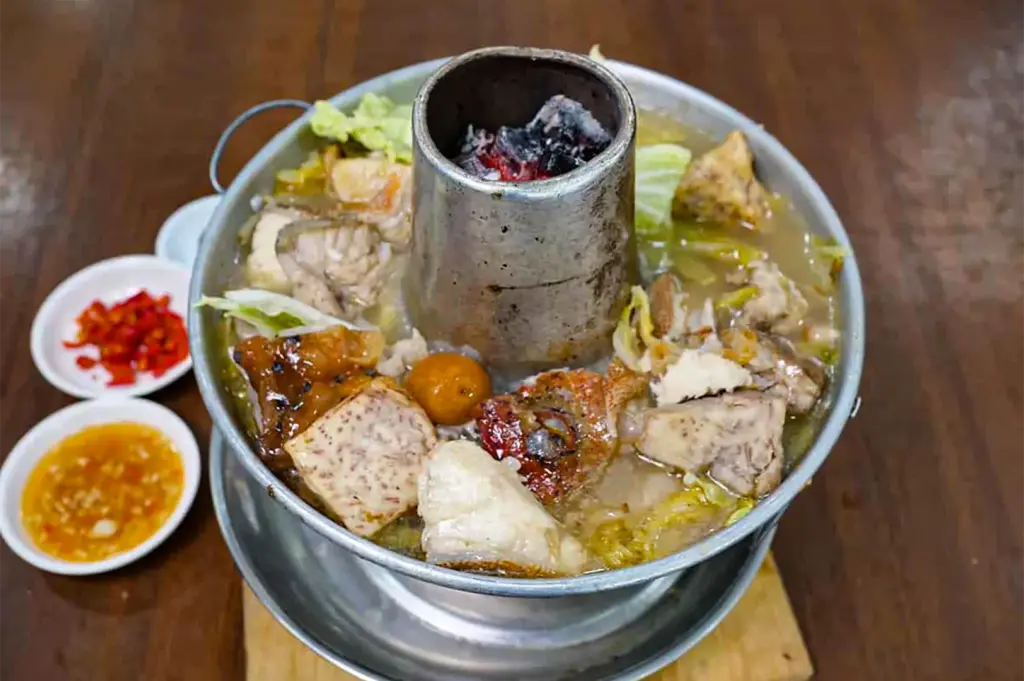 A traditional metal steamboat pot, powered by glowing charcoal in its central chimney, simmers with tender fish pieces, taro, and cabbage in a rich broth. Small dipping dishes of chopped red chilies and garlic sauce rest on the wooden table nearby.
