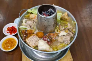 A traditional metal steamboat pot, powered by glowing charcoal in its central chimney, simmers with tender fish pieces, taro, and cabbage in a rich broth. Small dipping dishes of chopped red chilies and garlic sauce rest on the wooden table nearby.