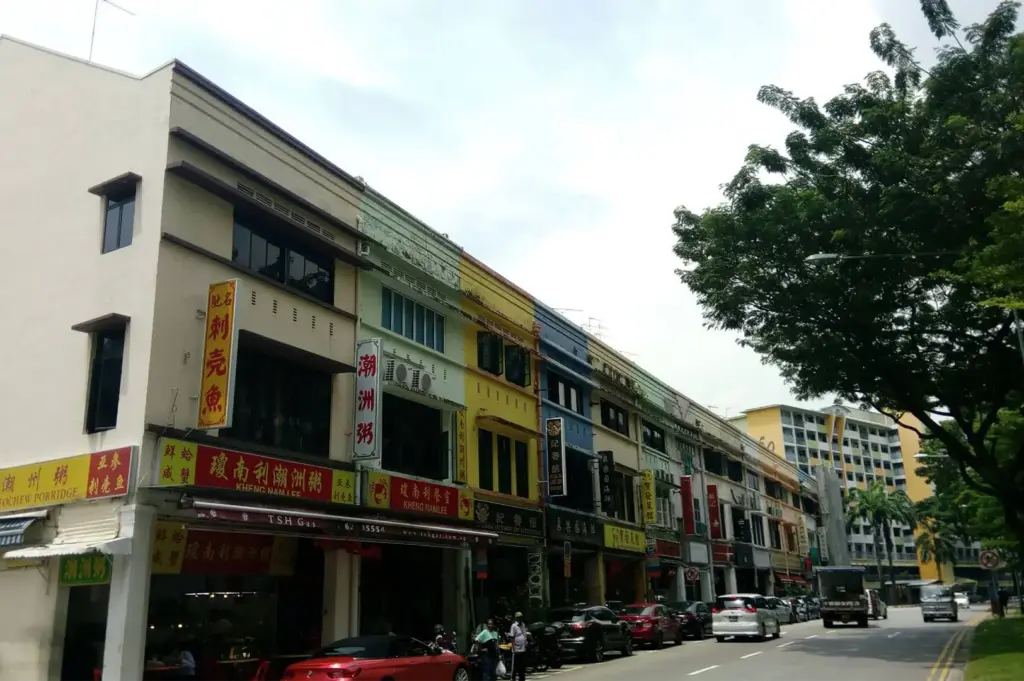 A wide exterior shot captures a row of colorful, multi-story traditional shophouses lining a city street with parked cars and leafy trees. Vibrant business signboards displaying Chinese characters and English text, including Teochew porridge eateries, mark the ground-floor shopfronts.