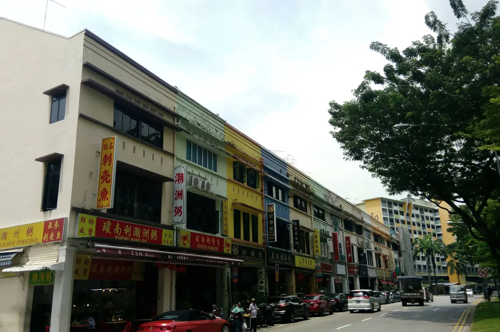 A wide exterior shot captures a row of colorful, multi-story traditional shophouses lining a city street with parked cars and leafy trees. Vibrant business signboards displaying Chinese characters and English text, including Teochew porridge eateries, mark the ground-floor shopfronts.