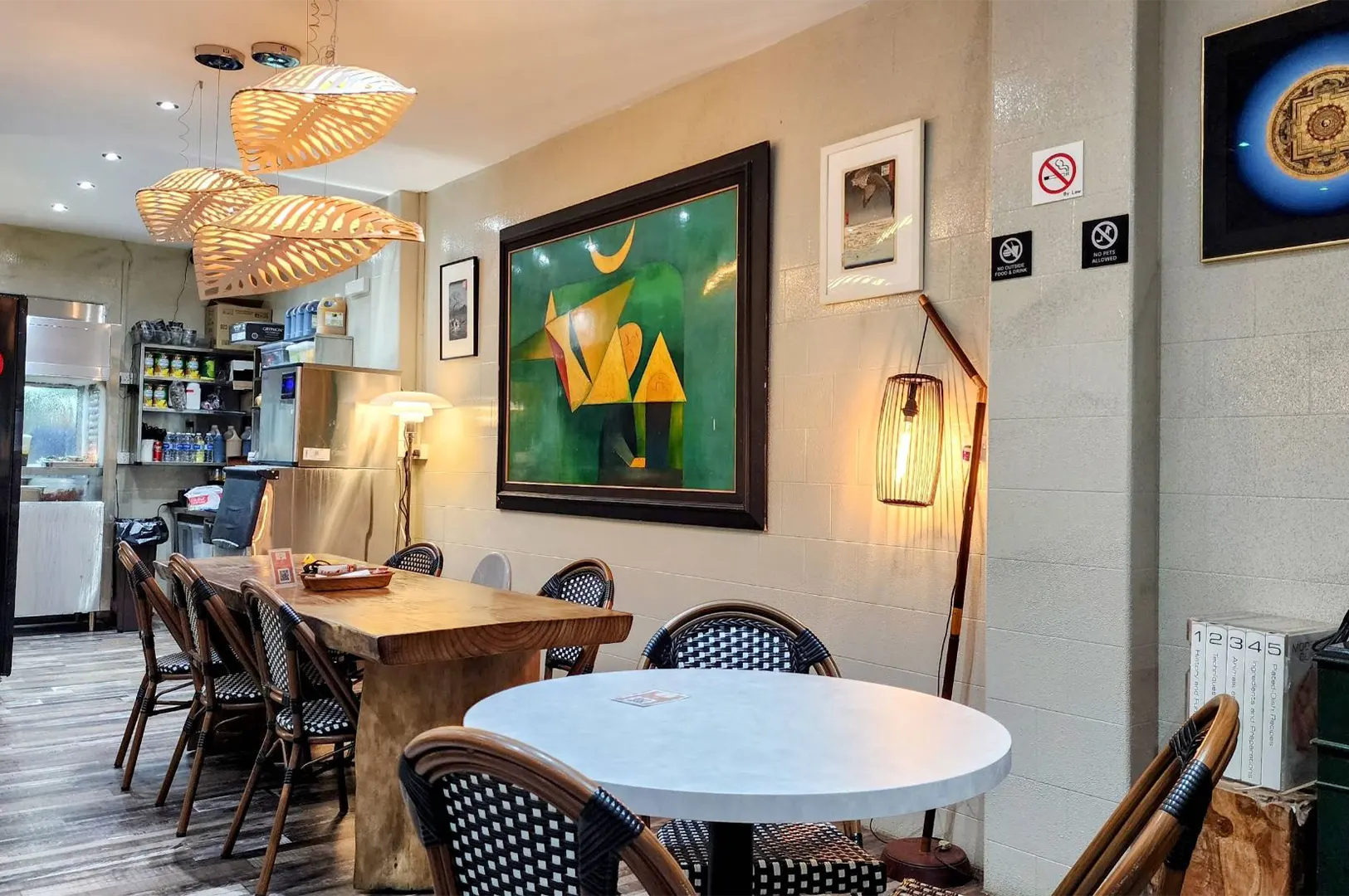 The interior of a warm, eclectic restaurant features round and rectangular wooden dining tables surrounded by woven rattan chairs. Warm illumination glows from leaf-shaped pendant lamps and a standing floor light, highlighting framed abstract artwork on the tiled wall.