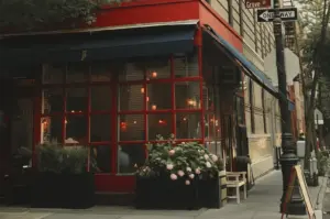 A charming corner bistro framed in bold red window trim and dark blue awnings glows with warm interior lighting on a quiet city street. Large planter boxes with lush pink hydrangeas decorate the sidewalk in front of the glass facade, near a "Grove St" street sign and black lamppost.