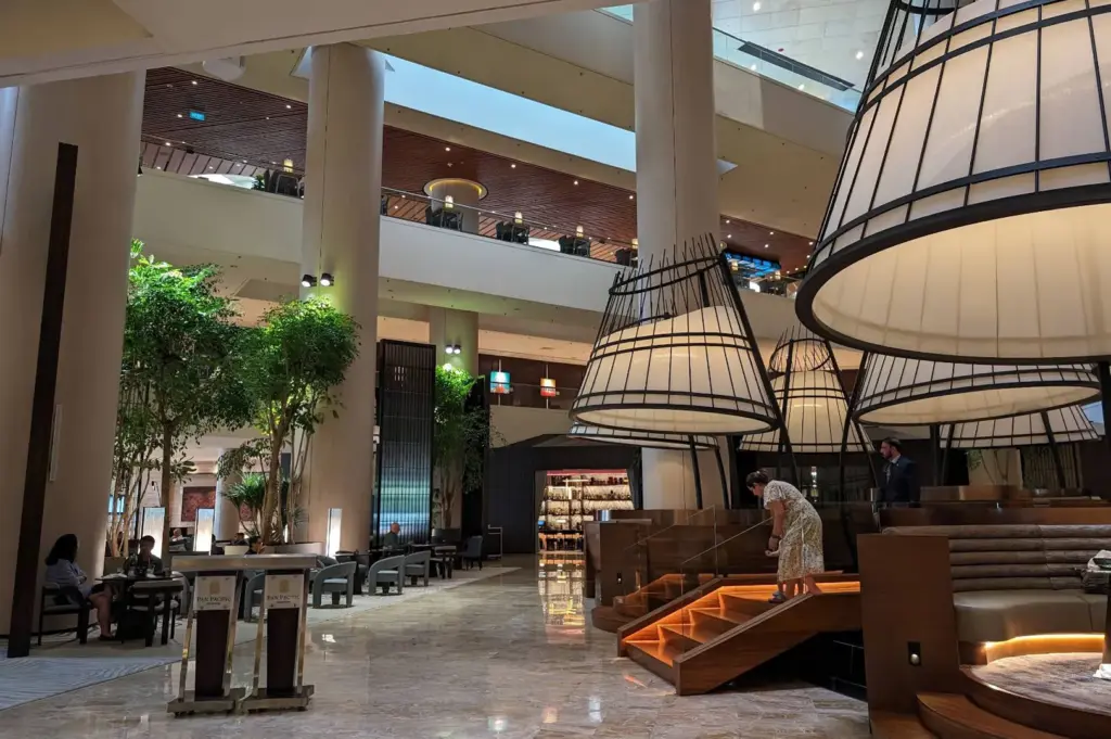 A soaring hotel lobby lounge features grand, lantern-inspired cage seating structures suspended above wooden elevated dining platforms. Multi-level balconies, tall concrete pillars, and lush indoor trees frame polished marble floors under soft architectural lighting.