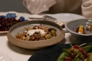 A shallow ceramic bowl featuring a gourmet pasta or ragù dish topped with dollops of white cream, grated cheese, and fresh microgreens takes center stage on a dinner table. Surrounding ceramic serving plates present artfully plated appetizers with roe, garnishes, and vibrant side dishes under soft, ambient lighting.