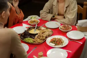 Friends gather around a red tablecloth spread with an abundant Mexican feast, featuring a central platter of crispy chicharrón, grilled meats, queso fundido, and guacamole. Side dishes of fresh pico de gallo, sliced cheese, and salsas are arranged beside individual white dining plates.
