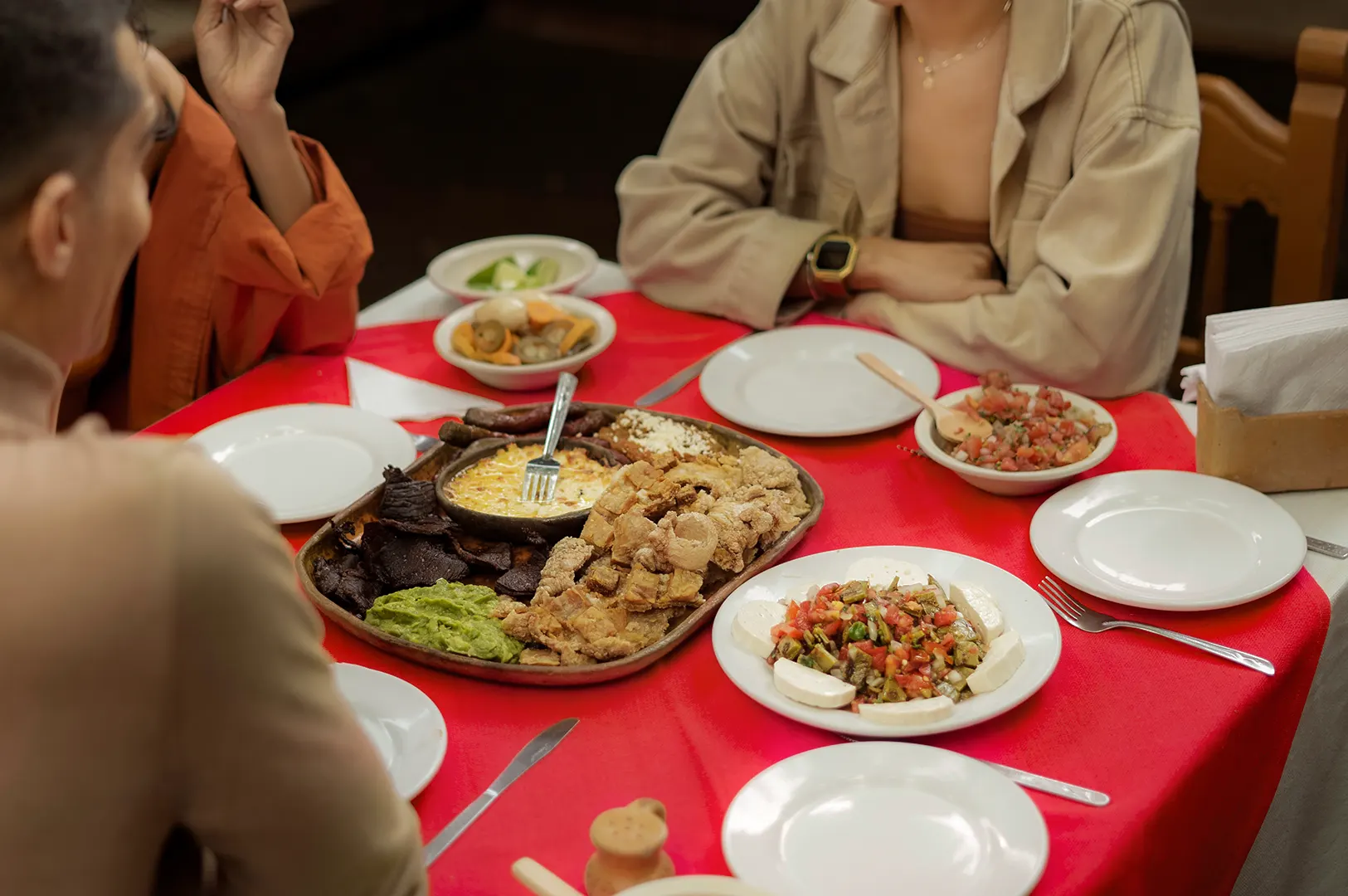 Friends gather around a red tablecloth spread with an abundant Mexican feast, featuring a central platter of crispy chicharrón, grilled meats, queso fundido, and guacamole. Side dishes of fresh pico de gallo, sliced cheese, and salsas are arranged beside individual white dining plates.
