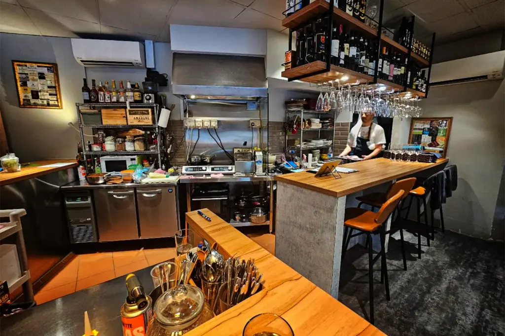 A wide-angle, eye-level shot captures the intimate, cozy interior of a small open-kitchen restaurant and bar, representing the file "1-Where Japanese Curry Meets Cozy.webp". On the right, a wooden service counter is paired with several tan leather barstools under an overhead rack loaded with wine bottles and rows of hanging glassware, where a chef wearing an apron stands ready to serve. The compact kitchen area in the center and left features stainless steel counters, a stovetop under a ventilation hood, storage racks filled with bottles and ingredients, a small microwave, and kitchen tools. In the immediate foreground, a wooden partition holds metal containers packed with spoons, forks, and other dining utensils.