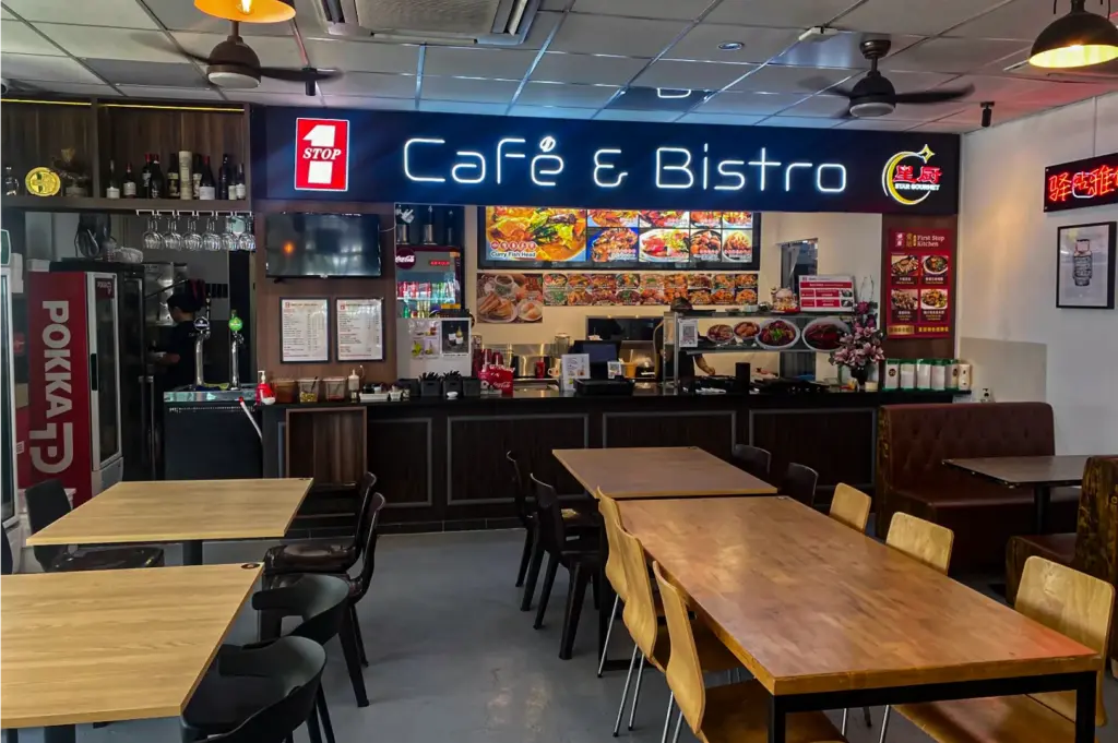 A illuminated blue neon sign reading "Café & Bistro" hangs above an ordering counter adorned with colorful food menu boards in a spacious dining area. Long wooden dining tables, wooden chairs, and plush booth seating fill the room beneath ceiling fans and warm pendant lights.