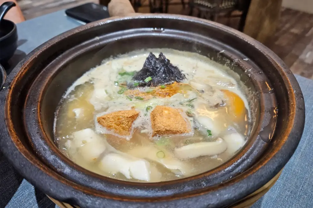 A steaming claypot fish soup features a rich, milky broth brimming with thick slices of pomfret, golden fried tofu puffs, sliced mushrooms, and a cluster of dark seaweed topped with scallions. The earthenware pot rests on a table, holding the freshly simmered soup hot and bubbling.