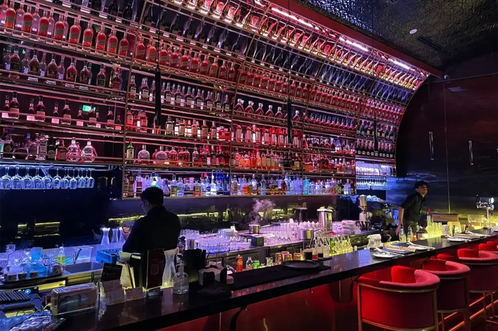 A stylish, dimly lit cocktail bar showcases a towering curved display shelf filled with rows of liquor bottles dramatically illuminated in vibrant pink and neon blue lighting. Bartenders work behind a long dark counter lined with plush red barstools, creating a chic, mood-lit atmosphere.