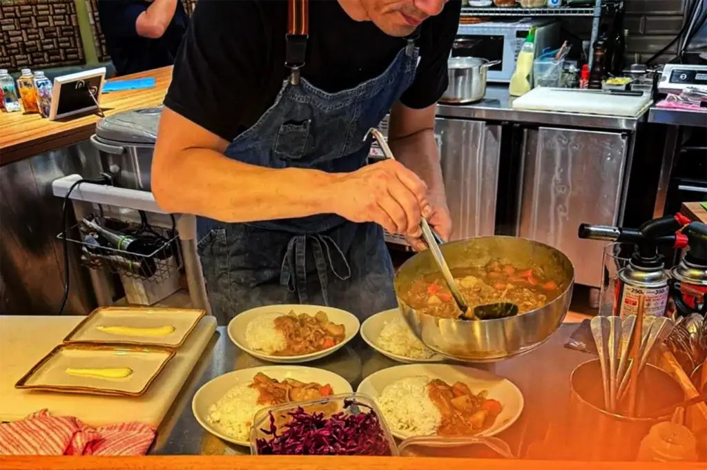 A close-up, medium shot captures a chef actively preparing multiple plates of food behind a service counter, representing the file "2-Where Japanese Curry Meets Cozy.webp". Wearing a black t-shirt and a blue denim apron, the chef stands leaning forward over a stainless steel prep station as he uses a large metal ladle to scoop thick, steaming Japanese curry from a large stainless steel pot onto four bowls filled with white rice. In the foreground, a container of shredded red cabbage sits ready for garnishing alongside two small rectangular plates smeared with a yellow sauce, while a kitchen blowtorch and canisters stand on the far right. The blurred commercial kitchen background features metal countertops, a microwave, and various cooking utensils.