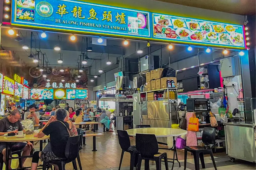 A vibrant, open-air food center features a bright blue backlit sign for "Hualong Fishhead Steamboat" alongside illustrated menu panels. Patrons sit at round dining tables under hanging ceiling lights, enjoying a lively, casual atmosphere surrounded by bustling food stalls.