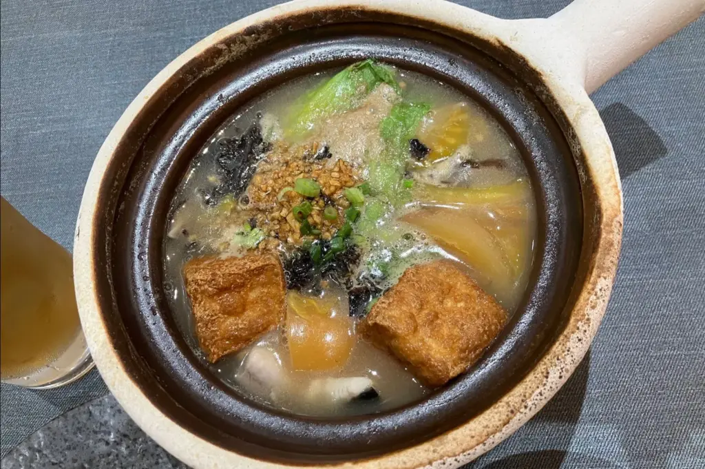 A top-down view shows a clay pot filled with hot, clear fish soup topped with golden-brown fried tofu puffs, fresh leafy greens, tomato wedges, and minced garlic garnish. The bubbling broth rests on a textured grey placemat alongside a tall glass of cold beverage.