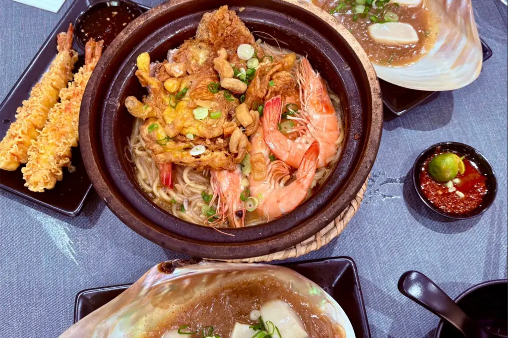 A claypot of seafood noodle soup takes center stage, filled with whole prawns, thick egg floss, scallions, and pork lard croutons. It is served alongside crunchy tempura prawns, chili dip with lime, and steamed scallops presented in large, iridescent seashells.