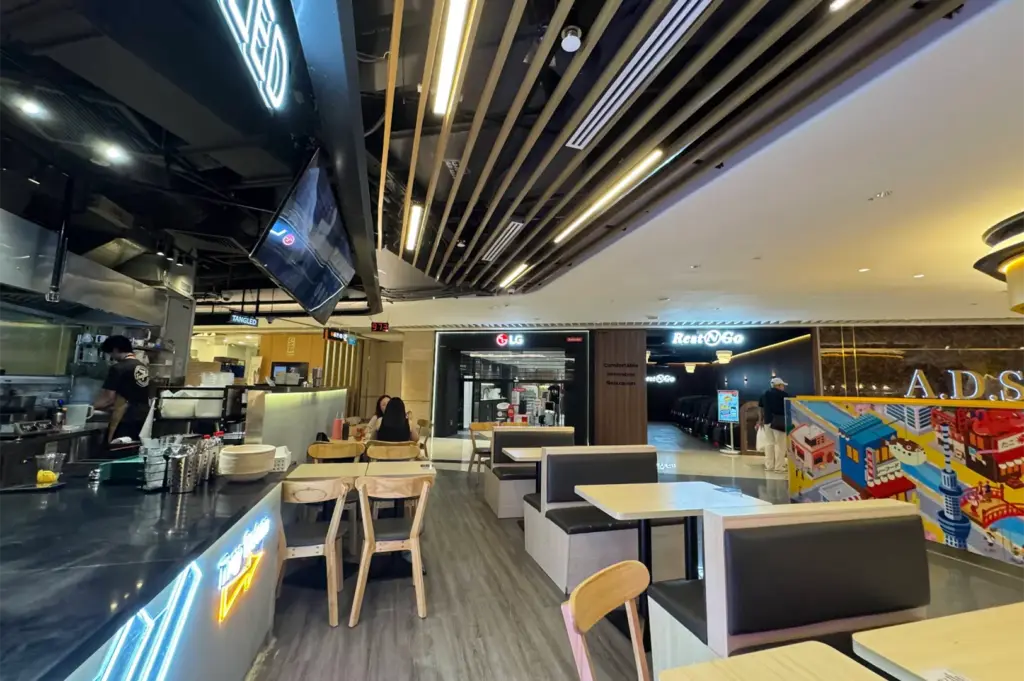 A modern indoor food court feature dining area booth seating, light wooden chairs, and an open kitchen counter accented with neon light fixtures. Slatted wooden ceiling panels extend toward adjacent retail spaces, including an LG store and a massage chair lounge.