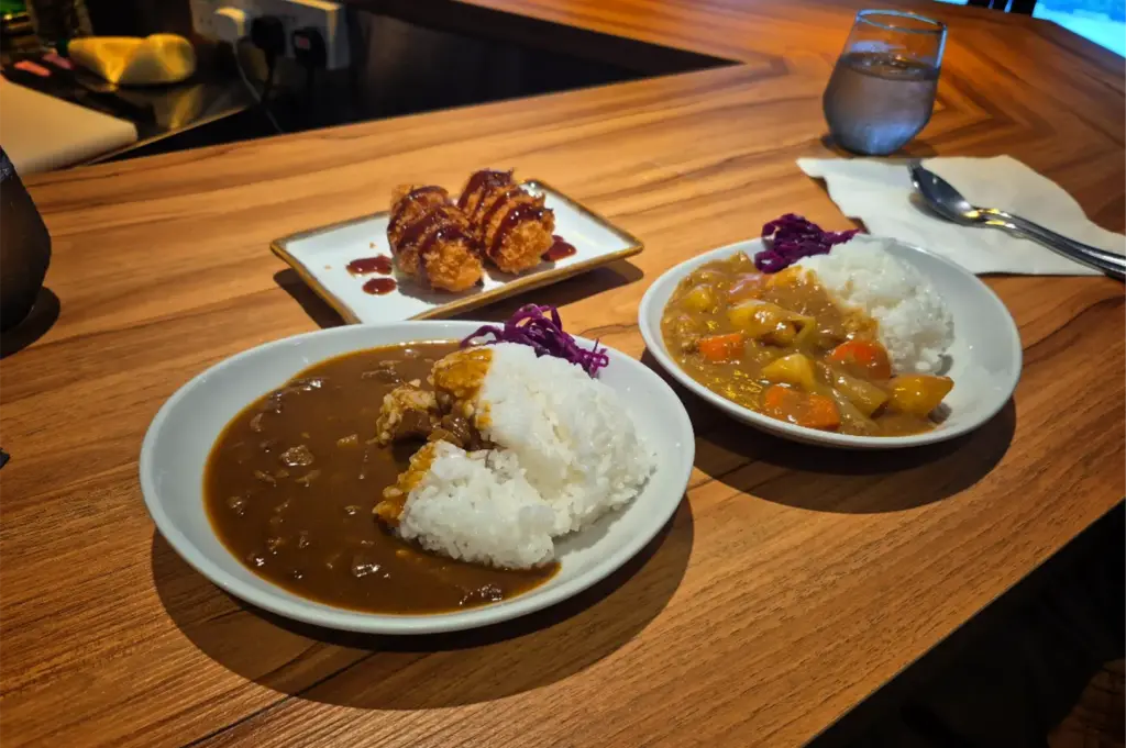 A high-angle, medium shot captures a neatly arranged Japanese meal served on a warm wooden countertop, representing the file "3-Where Japanese Curry Meets Cozy.webp". In the foreground, a white oval plate features a mountain of white rice split down the middle by a rich, dark brown beef curry sauce, garnished with a small heap of shredded red cabbage. Just behind it to the right sits a second plate of lighter-colored curry filled with visible chunks of carrots and potatoes next to a serving of rice, while a rectangular side plate to the left holds two golden, deep-fried katsu cutlets drizzled with a dark tonkatsu sauce. In the upper right corner, a glass of water, a spoon, and a fork resting on a white napkin complete the cozy dining setup.