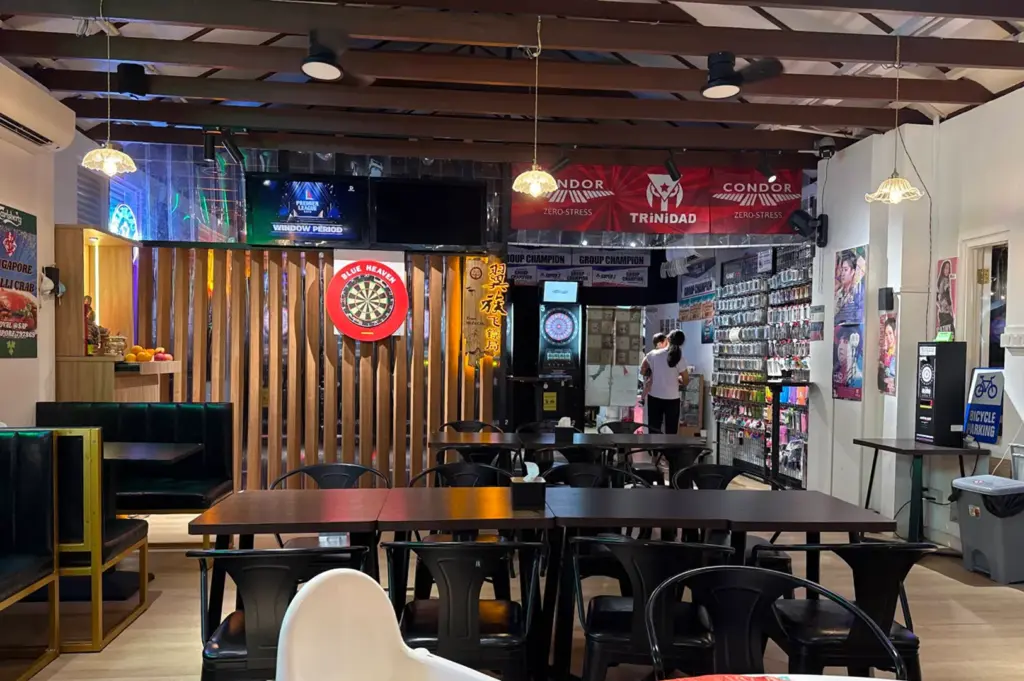 A lively sports bar and eatery features dark dining tables and black metal chairs beneath exposed wooden ceiling beams and vintage pendant lamps. Mounted dartboards, branded banners, TV screens, and a retail display wall add energy to the spacious indoor venue.