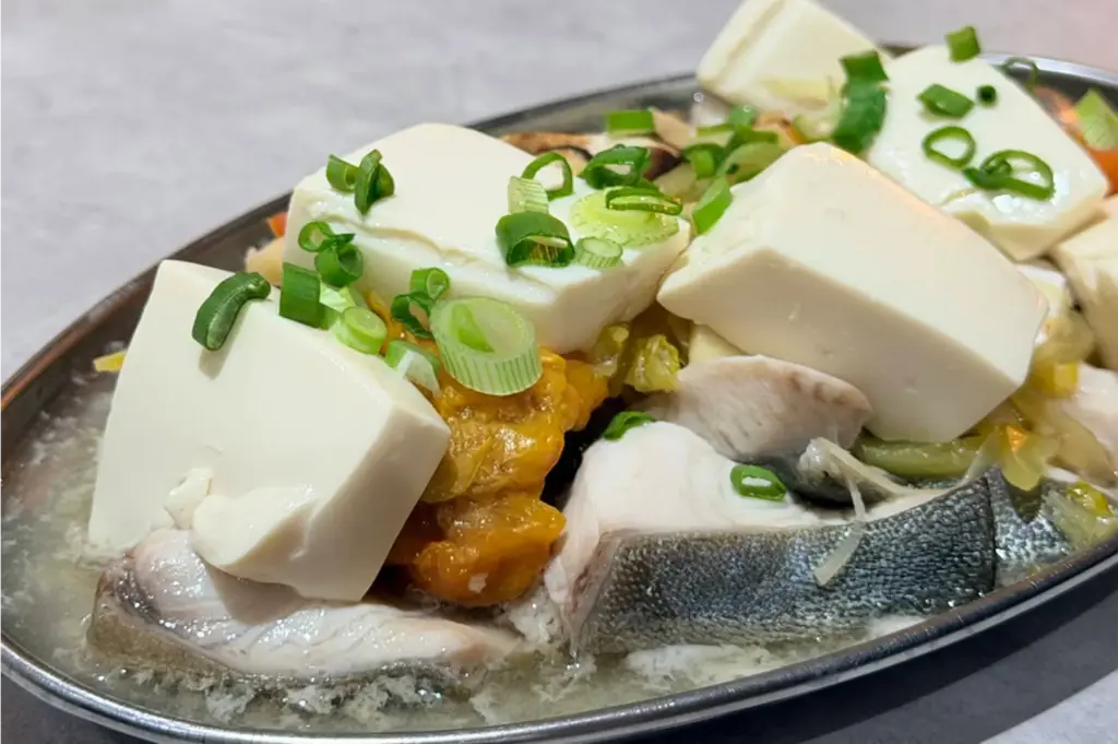 A close-up shot displays a steamed fish dish served on an oval stainless steel platter, featured with delicate fish fillets, silky white tofu blocks, and salted vegetables. The dish is garnished with sliced scallions and bathed in a light, savory broth.