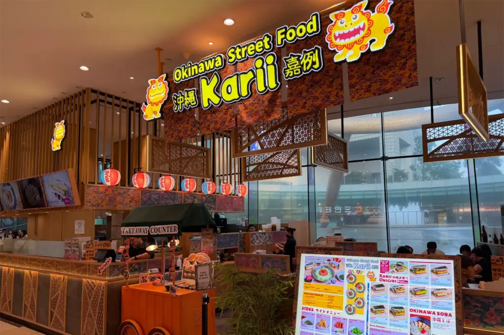 A brightly lit Japanese eatery features bold signage for "Okinawa Street Food Karii" adorned with vibrant shisa lion illustrations, patterned fabric banners, and Orion paper lanterns. An orange takeaway cart stands near a menu board at the front, with expansive floor-to-ceiling windows offering a view of an outdoor fountain in the background.