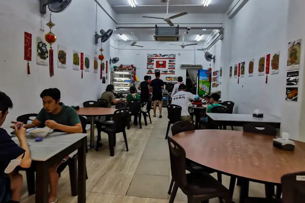 A brightly lit, casual dining hall features wooden round and square tables surrounded by dark plastic chairs where patrons enjoy their meals. White walls decorated with red traditional Chinese ornaments and food posters line the narrow space, framed by ceiling fans overhead.