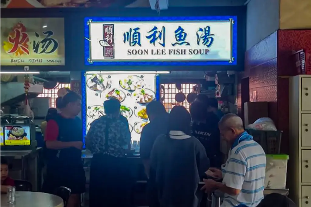 A illuminated signage reading "SOON LEE FISH SOUP" in English and Chinese characters marks a busy food stall in a hawker center. Customers queue up in front of the counter, which displays a illuminated photo menu of various fish soup dishes.