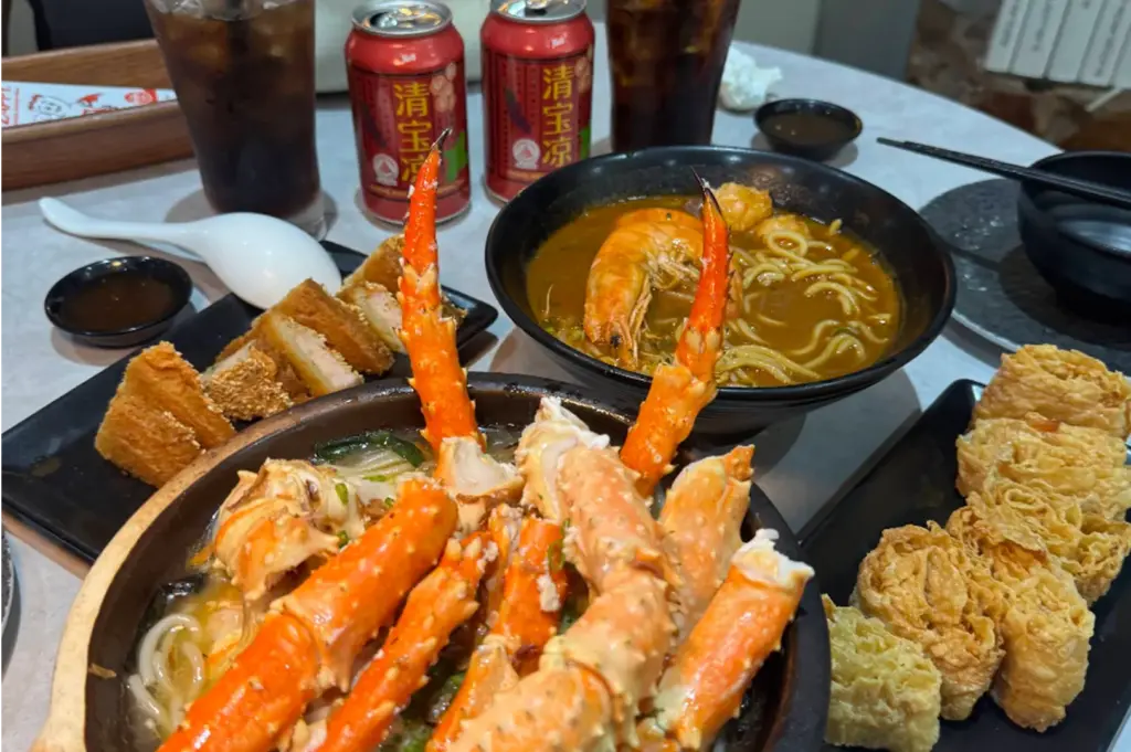 A spread of Asian seafood dishes features a claypot bowl overflowing with bright orange crab legs alongside a bowl of prawn noodle soup. Crispy fried side dishes, iced drinks, and canned herbal tea surround the main bowls on the dining table.