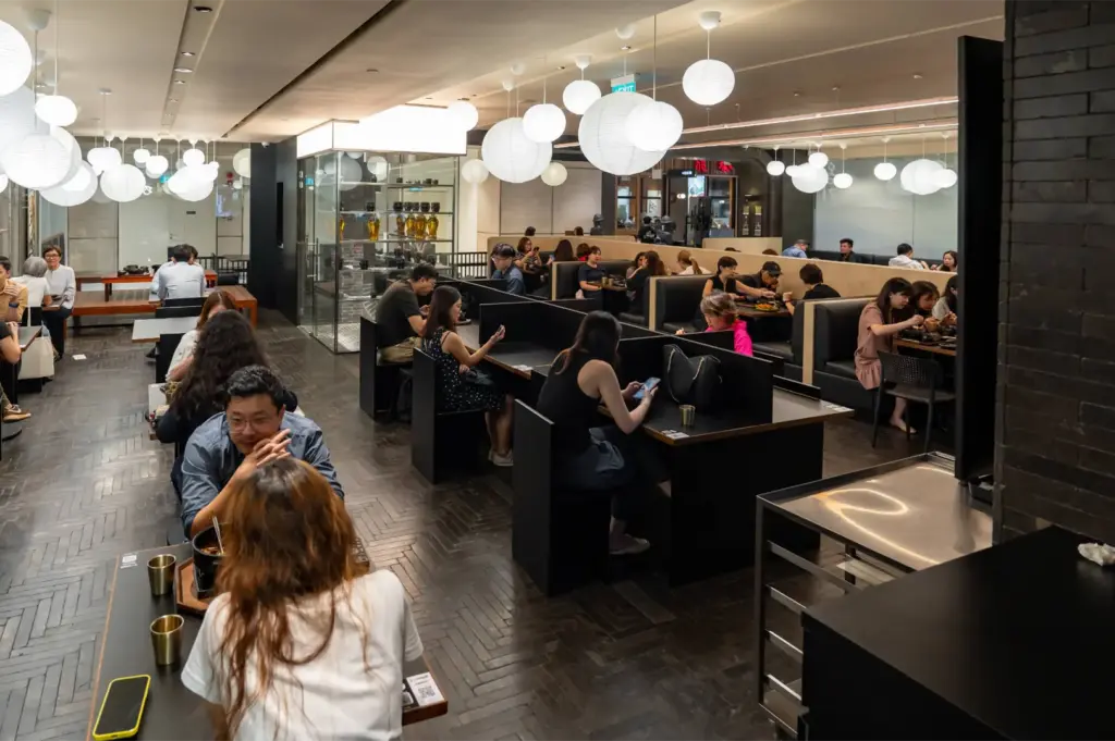 A spacious, bustling dining room features dark herringbone floors and numerous white paper lantern lights suspended from the ceiling. Diners occupy dark wood booth seating and individual partitions, creating a lively yet organized atmosphere throughout the restaurant.
