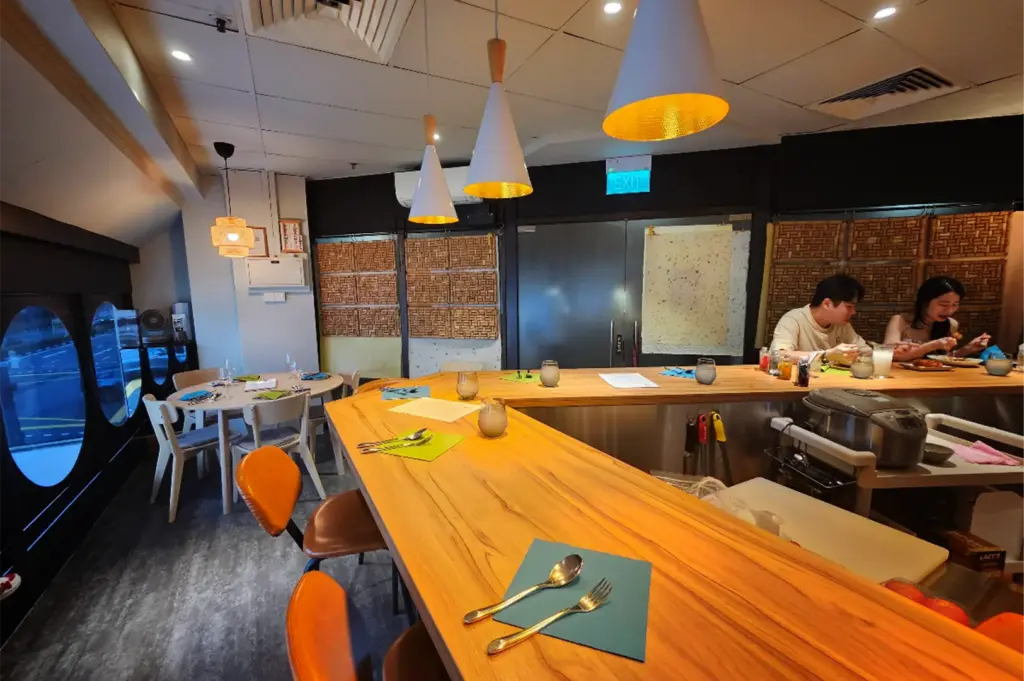 A wide-angle, eye-level shot captures a modern and warmly lit restaurant dining room, representing the file "5-Where Japanese Curry Meets Cozy.webp". A long, angular wooden bar counter dominates the foreground and center, lined with a few orange-and-brown leather barstools, set utensils on colorful napkins, and drinking glasses. In the background on the right, a couple sits at the far end of the counter enjoying a meal near a kitchen prep station, while a separate round dining table with empty chairs sits near distinct circular windows on the left, looking out onto a road. Overhead, a trio of conical pendant lights with reflective gold interiors illuminates the wooden counter, casting a warm glow across the cozy interior.