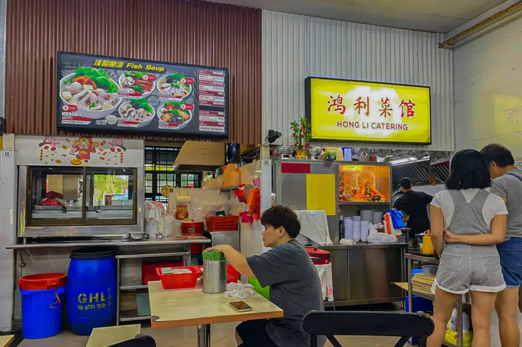 A bustling hawker stall counter features prominent signage for "Hong Li Catering" and an illuminated menu board highlighting various fish soup dishes. Customers sit at nearby tables and wait near stainless steel food preparation areas against corrugated wall paneling.
