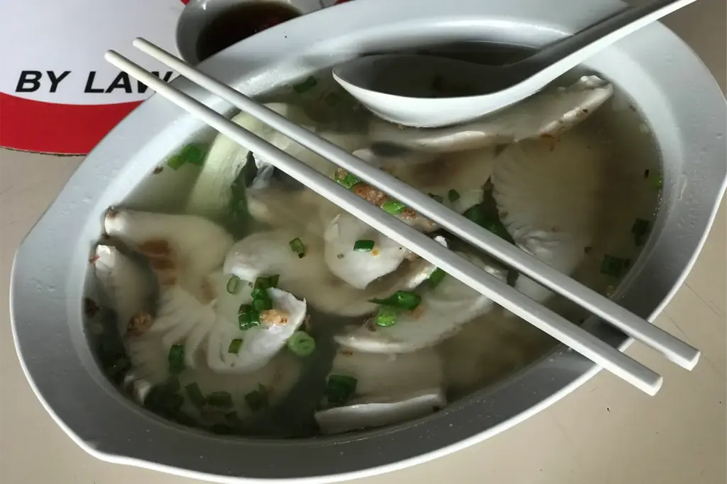 A large oval bowl is filled to the brim with clear fish soup loaded with sliced white fish fillets and sprinkled with finely chopped scallions. A white ceramic soup spoon and a pair of white chopsticks rest balanced across the top rim of the bowl.