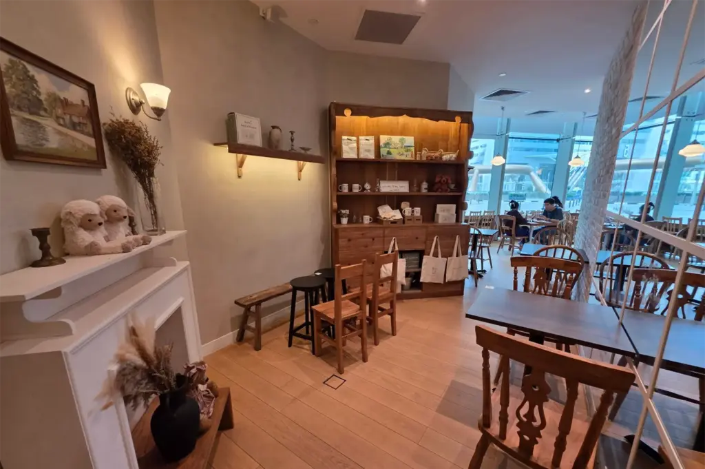 A cozy cafe interior features warm wooden furniture, rustic shelving display units, and a faux fireplace adorned with plush lamb toys and dried botanical arrangements. Soft lighting accents beige walls and light wood flooring, while floor-to-ceiling windows bring natural light into the background dining area.