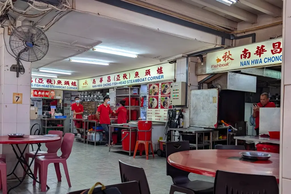 An open-concept restaurant dining room features prominent signage for "Nan Hwa Chong Fish-Head Steamboat Corner" above kitchen service areas. Staff members wearing matching red shirts stand near stainless steel prep stations, with round plastic dining tables arranged across the tiled floor beneath ceiling fans and bright fluorescent lights.