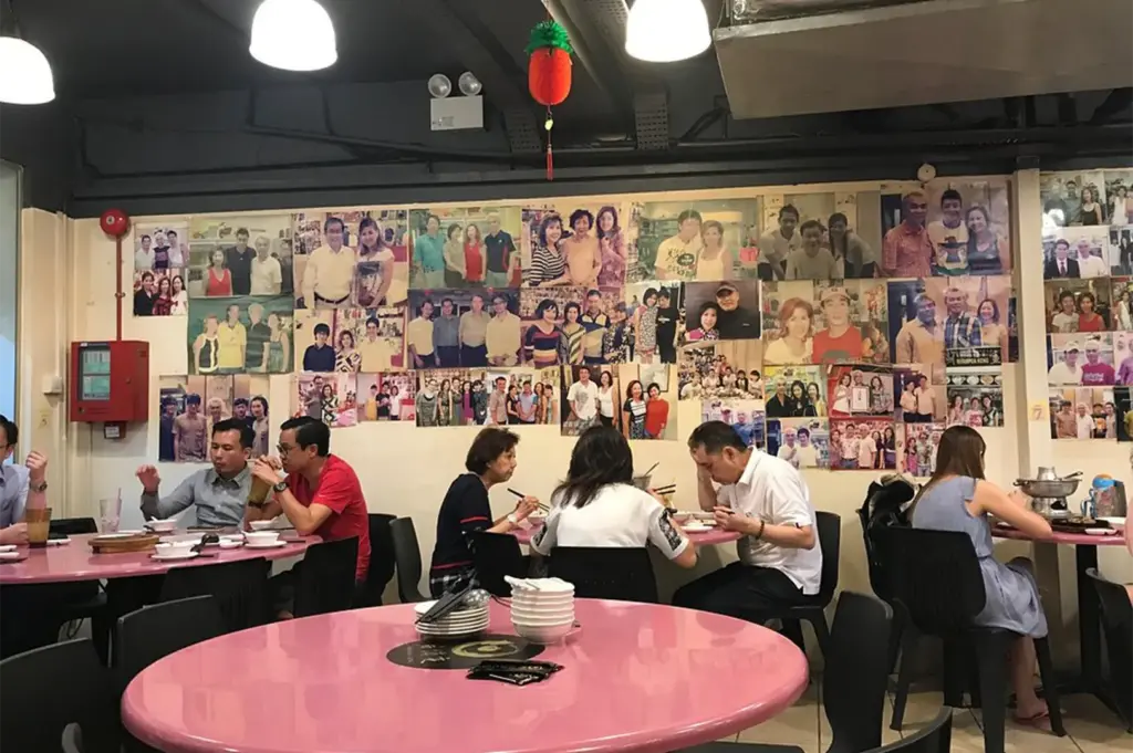 A lively dining room is filled with patrons seated around glossy pink round tables set with white dishware and small hot pots. Hanging dome lights illuminate a feature wall completely covered in a collage of printed photos capturing past diners and special moments.